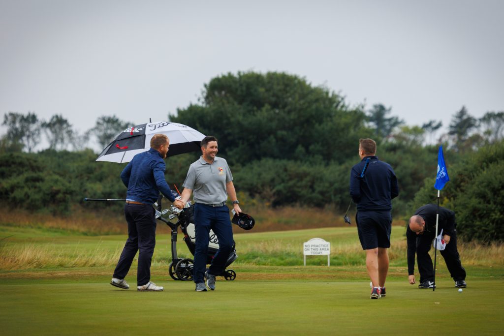 Golfers shake hands at the end of the Team Matchplay at the Men's Area Team Championship Final at Powfoot Golf Club in August 2022. There are three golfers, all male, and all smiling at each other. 